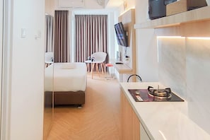 A hotel room with a bed, a small table with a chair, a television mounted on the wall, and a kitchen area with a stove. - Practical Studio At Tamansari Skylounge Makassar Apartment (Makassar)