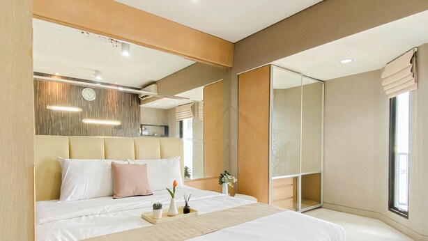A modern hotel room with a large bed, a bedside table with a vase and a plant, a wall-mounted clock, and a large window with curtains. - 2 Pax Serene Studio At Tamansari Sudirman Apartment (Jakarta)