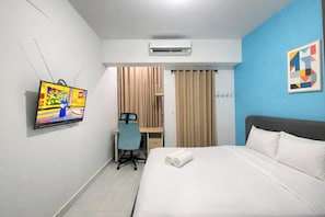 Habitación de hotel con cama, escritorio y silla, aire acondicionado, televisor y pared azul con un cuadro geométrico.