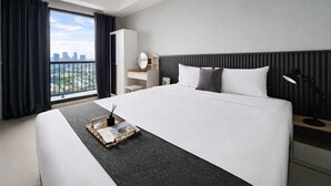 A hotel room with a large bed, a nightstand, a desk, a mirror, and a window with a city view. - Cozy And Minimalist 2Br At The Ayoma Apartment (South Tangerang)