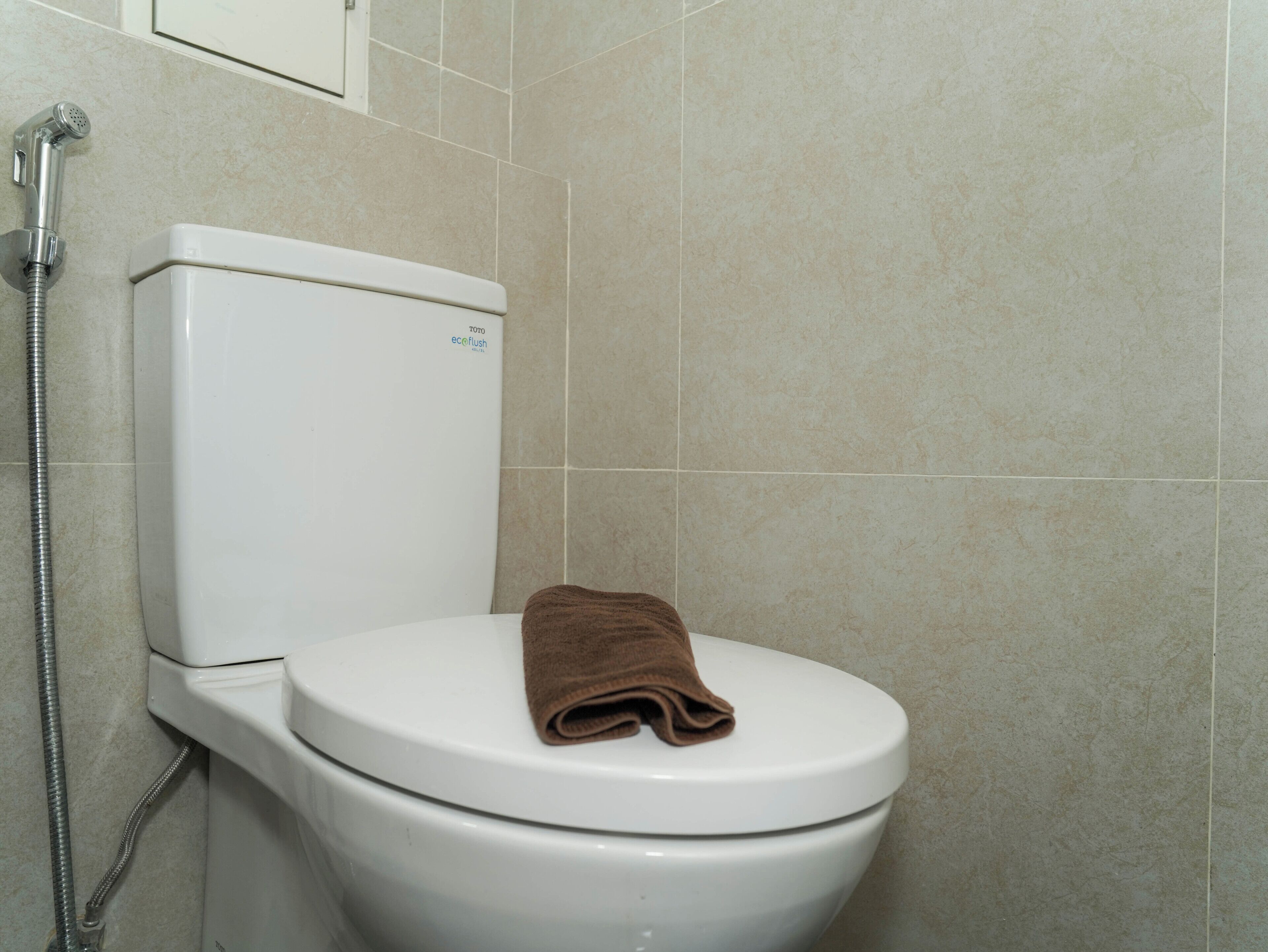 Inodoro blanco con una toalla marrón sobre la tapa, una manguera de ducha y paredes con azulejos color beige.