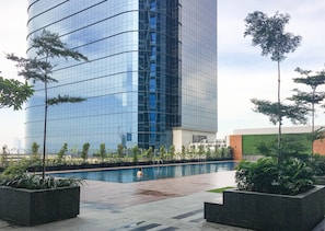 A modern building with a reflective glass facade, a swimming pool, and surrounding greenery. - Good Deal Studio With Modern Classic Style At The Vertu Apartment (Surabaya)