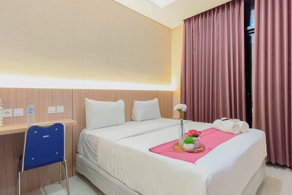 A hotel room with a bed, a desk, a chair, a water bottle, a vase with flowers, and a rolled towel. - Good Deal Studio With Modern Classic Style At The Vertu Apartment (Surabaya)