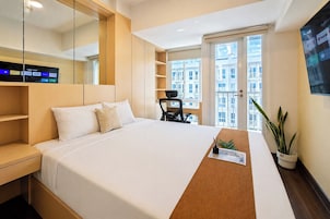 A modern hotel room with a large bed, a desk, a chair, a potted plant, and a TV displaying streaming services.