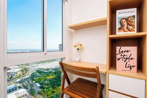 Quarto com uma janela grande, uma escrivaninha de madeira, uma cadeira, um vaso com flores e estantes repletas de livros.