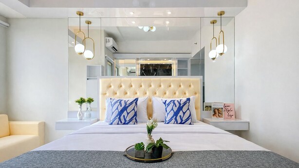 A modern bedroom with a tufted headboard, a bed with white bedding, two blue patterned pillows, a bedside table with a book, and a small plant on a tray. - Comfy White Studio At Transpark Juanda Bekasi Timur Apartment (Bekasi)