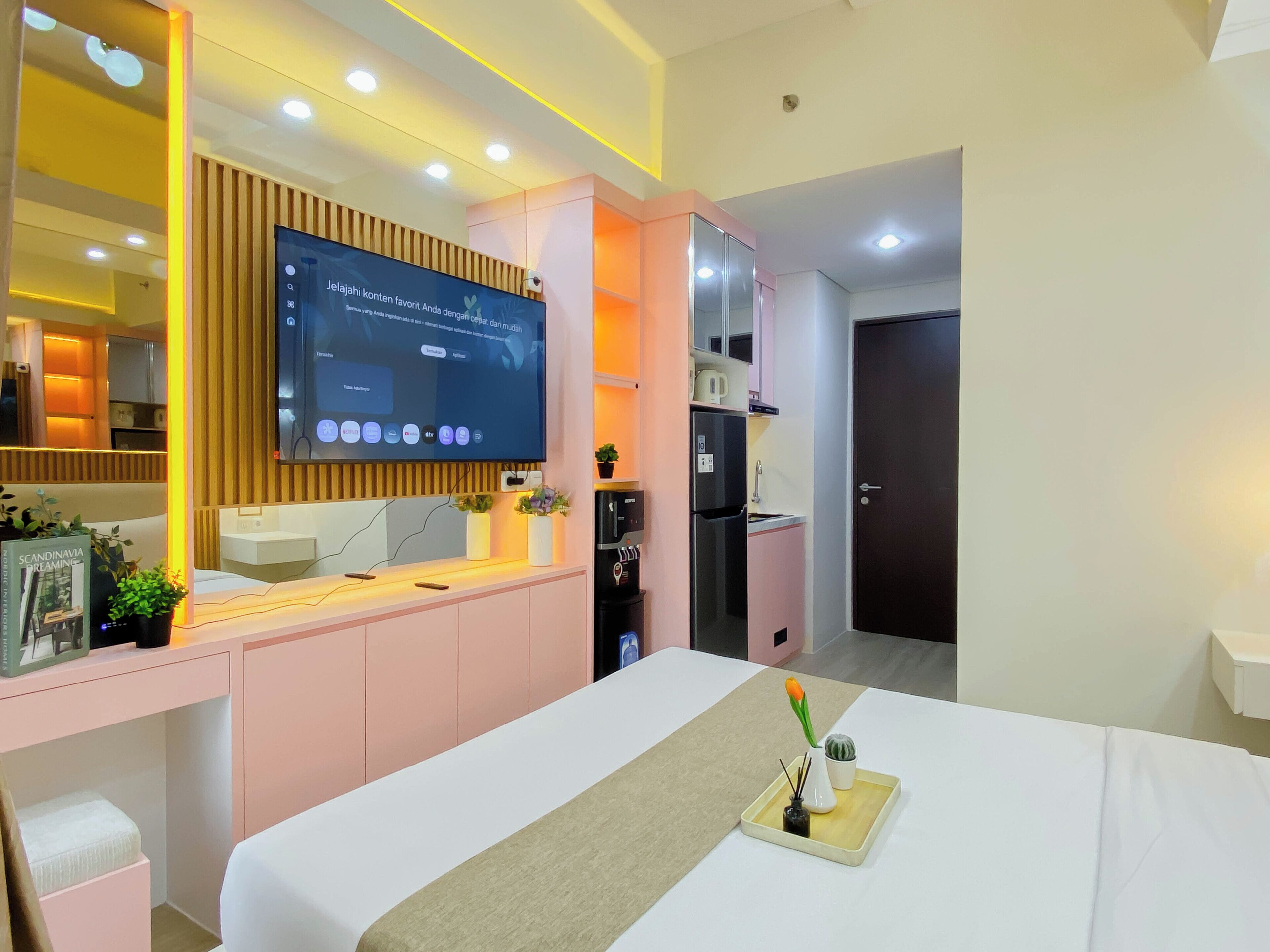 A modern hotel room with a bed, a TV mounted on the wall, a kitchenette, and a door.