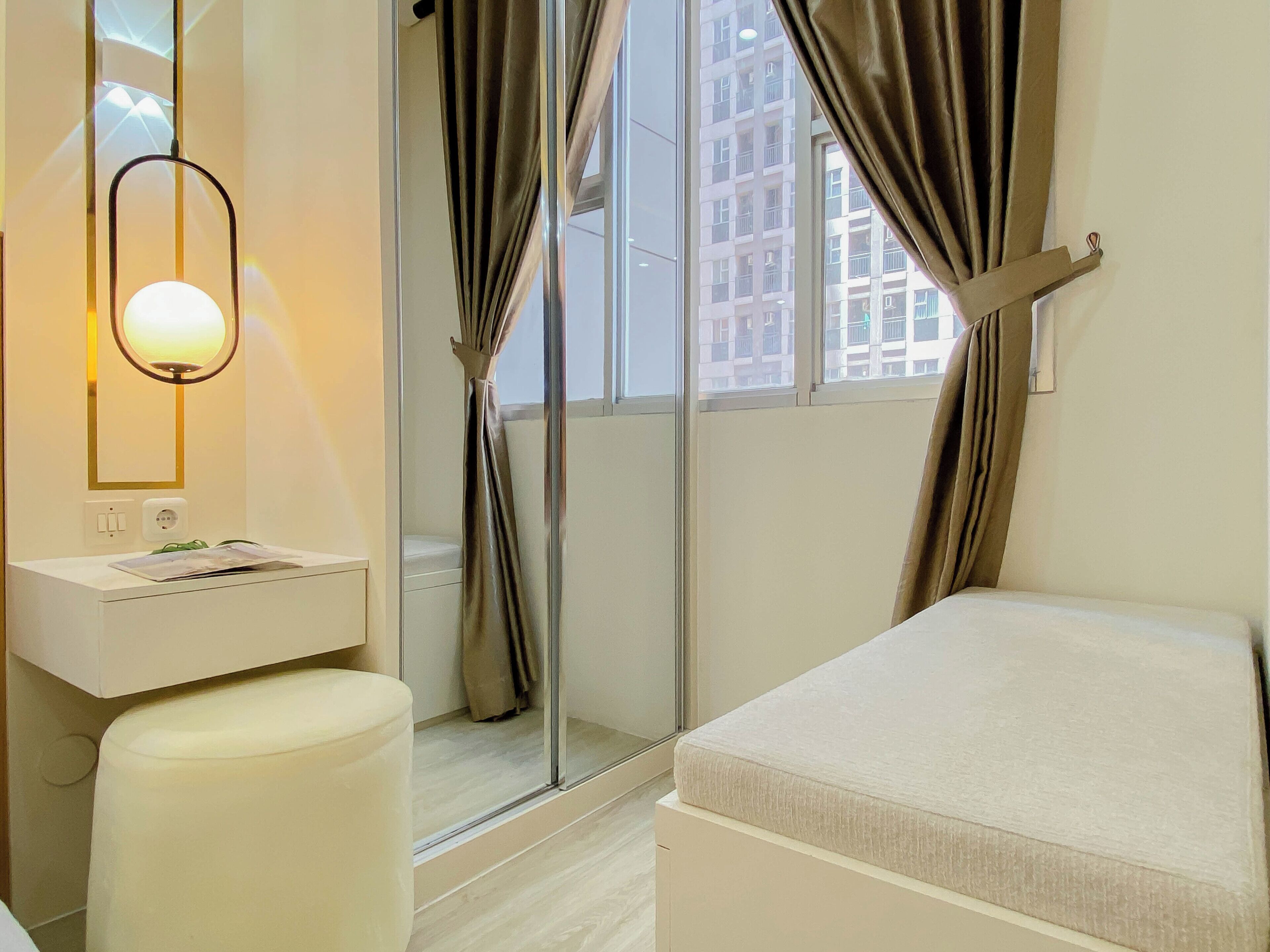 A modern hotel room with a bathroom, a bedside table, a window with curtains, and a view of buildings outside.