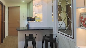 A modern kitchen with a black countertop, two black bar stools, a window with a diamond pattern, and a yellow appliance. - Functional And Cozy 2Br Apartment At Vida View Makassar (Makassar)