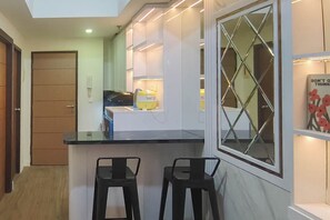 A modern kitchen with a black countertop, two black bar stools, a window with a diamond pattern, and a yellow appliance. - Functional And Cozy 2Br Apartment At Vida View Makassar (Makassar)