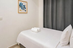 Una habitación de hotel con una cama, una toalla enrollada, un almohadón, una obra de arte enmarco y una cortina gris.