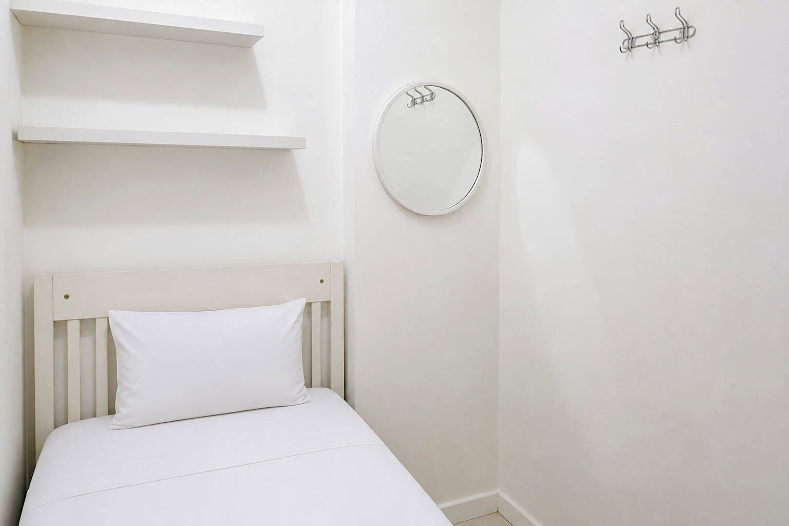 A white bed with a pillow, a round mirror, and a coat rack on a plain wall.