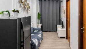 A compact living space with a refrigerator, a bed with a floral bedspread, a small table with a plant, and a grey curtain. - Cozy Living 2Br At Vida View Makassar Apartment (Makassar)