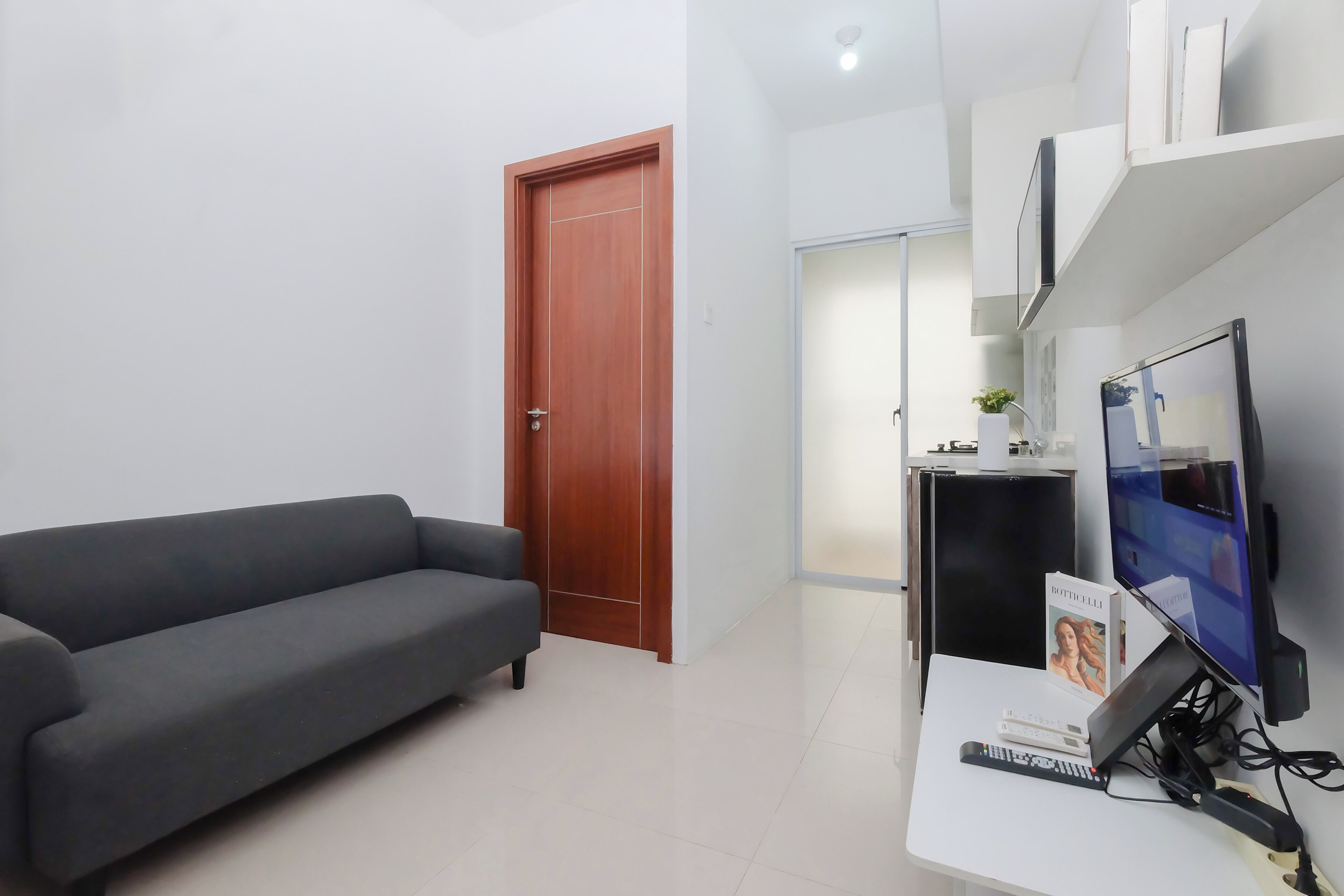 A modern living room with a grey sofa, a wooden door, a white desk with a computer and remote control, and a TV.