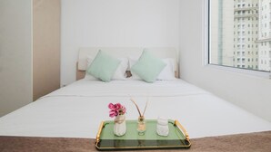 A neatly made bed with white bedding and two green pillows, a small table with a tray holding a vase with pink flowers, a glass bottle, and a small white figurine. - Tranquil 1Br For 2 Pax At Vittoria Residence (Jakarta)