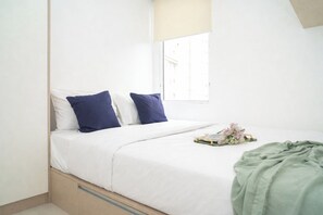 Ein Bett mit weißer Bettwäsche, zwei blaue Kissen, einer grünen Decke und ein kleiner Tisch mit einem Buch und Blumen.