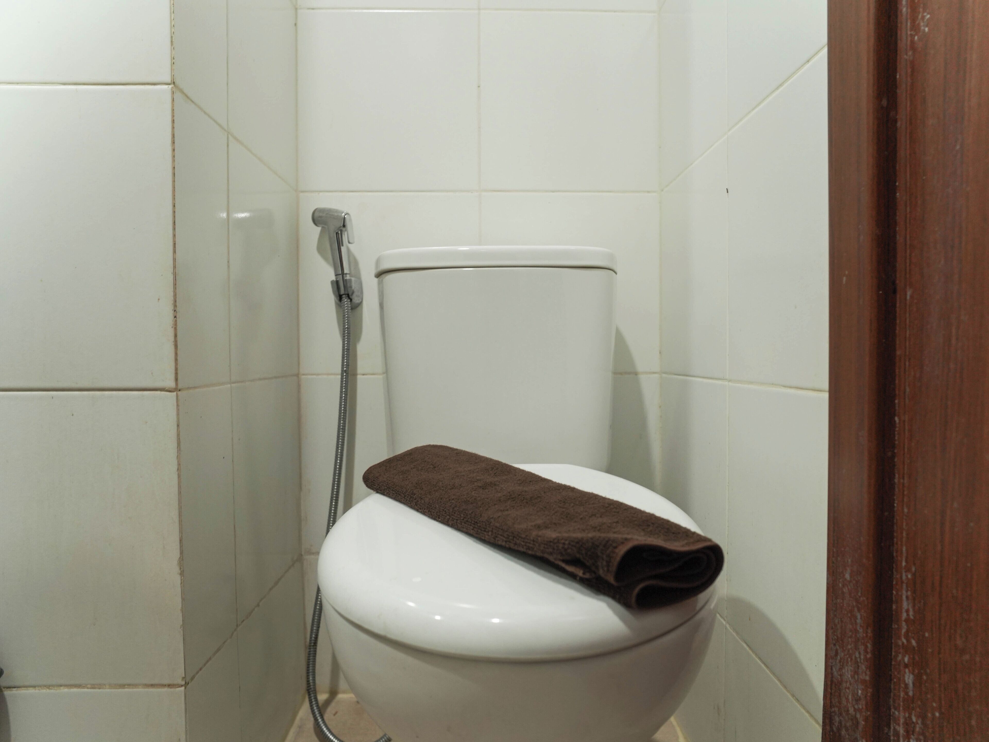 A white toilet with a brown towel on the seat, a shower hose, and white tiled walls.