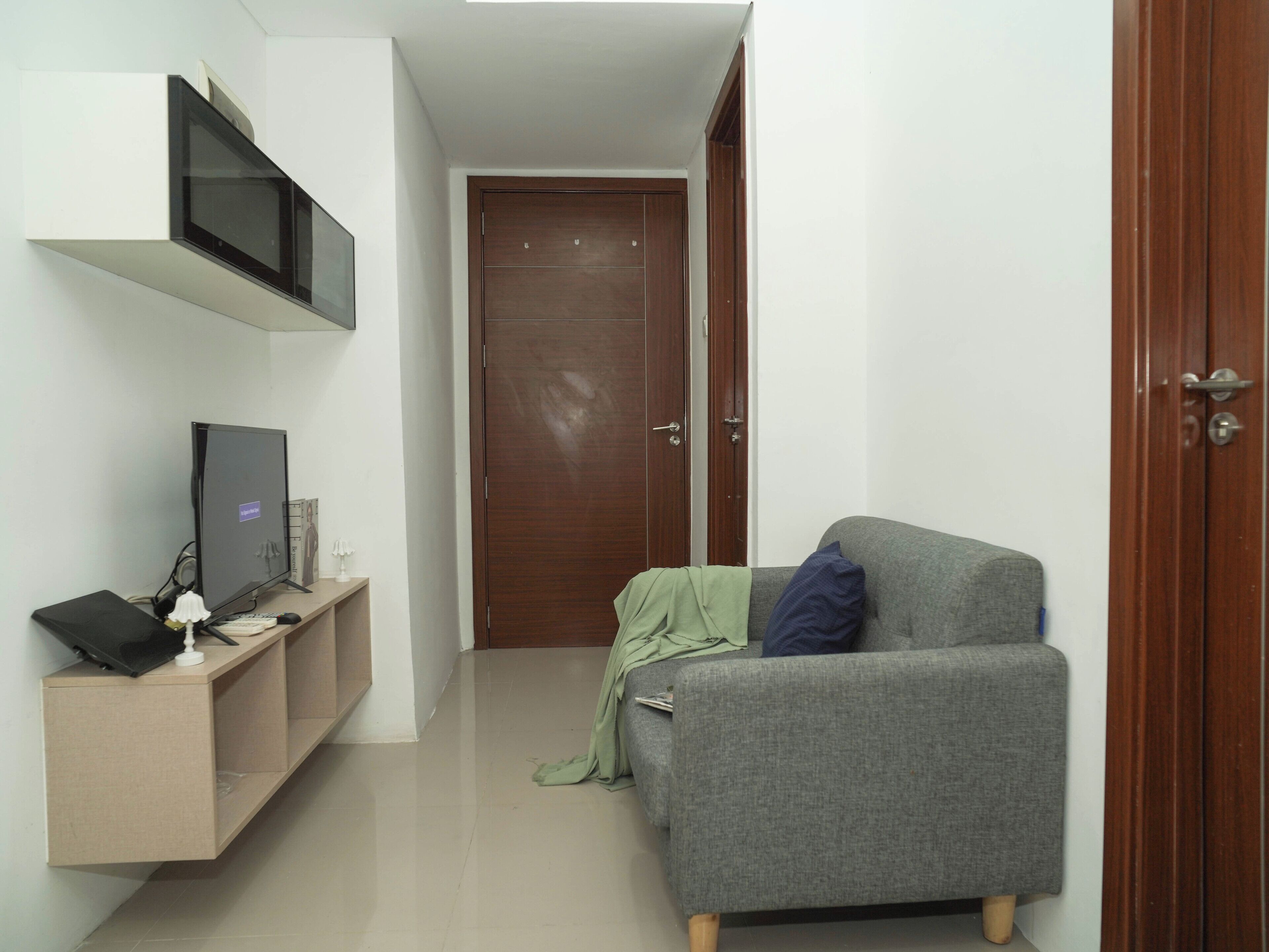 A small, narrow room with a grey sofa, a television on a stand, and a wooden door.