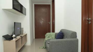 A small, narrow room with a grey sofa, a television on a stand, and a wooden door.