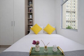 A neatly arranged bedroom with a bed, a side table, a bookshelf, and a window with a view of a building. - 3 Pax Tranquil 2Br At Vittoria Residence (Jakarta)