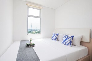 Ein ordentlich eingerichtetes Schlafzimmer mit einem Bett, Kissen, einem kleinen Tisch mit einer Vase und einem Fenster mit Blick auf Gebäude.