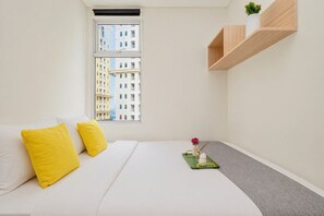 A hotel room with a bed, two pillows, a window with a view of buildings, a shelf with a plant, and a tray with a flower and candle. - Functional 2Br Apartment At Vittoria Residence (Jakarta)