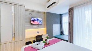 Ein Hotelzimmer mit Bett, einem an der Wand befestigten Fernseher, einem Schreibtisch mit Stuhl, einem Fenster mit Vorhängen und einer Klimaanlage.