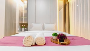 A hotel room with a bed, pillows, a pink bedspread, a rolled towel, a small potted plant, and a tray with a plant. - Stylish Light Gray Touch Studio At Westown View Surabaya Apartment (Surabaya)