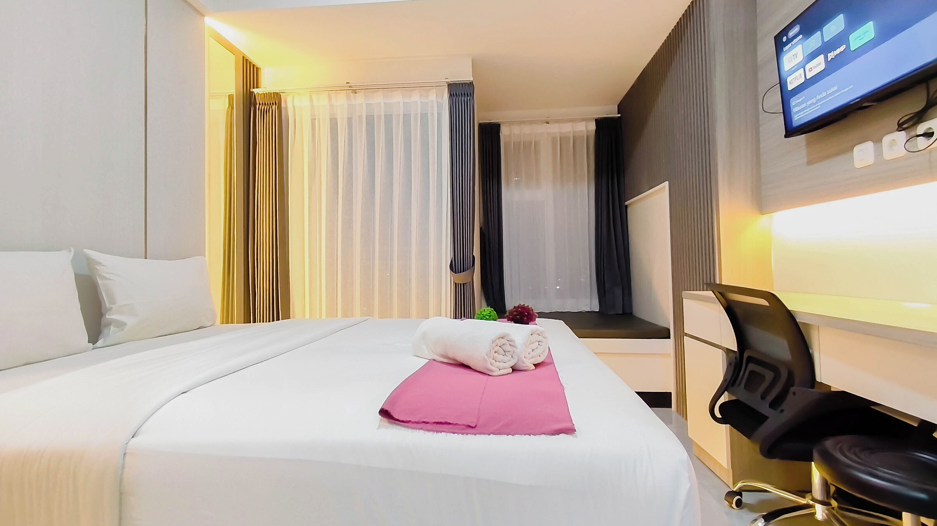 Quarto de hotel com cama, escrivaninha com cadeira, televisão e cortinas.