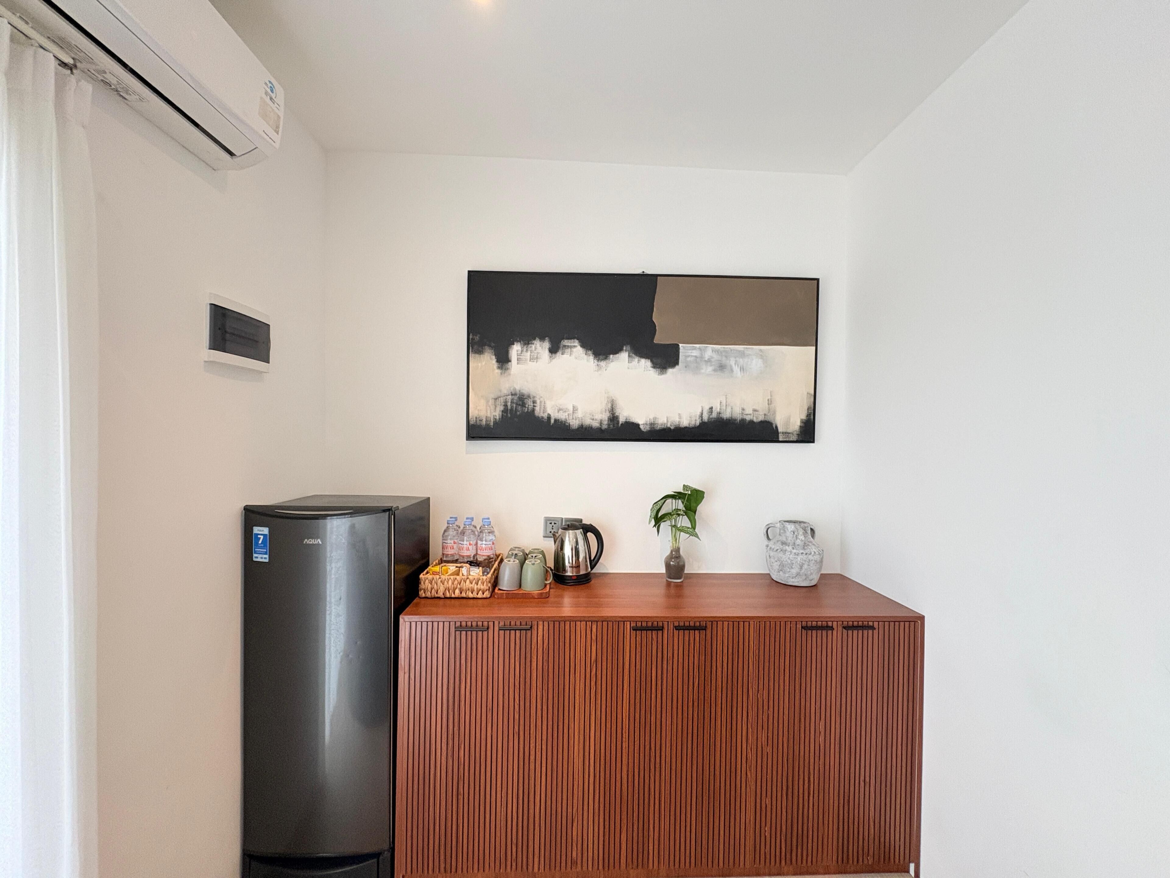A kitchenette with a refrigerator, a coffee maker, a kettle, and a plant on a wooden cabinet.