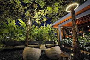 Un área de estar al aire libre con mobiliario de mimbre, rodeada de exuberante vegetación y una estructura de madera.