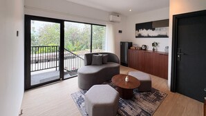 A modern living room with a sofa, ottoman, and coffee table, a sliding glass door to an outdoor balcony, and a kitchen area with a refrigerator and cabinets. - Guru Giri Forest Cabin Villa 2BR for 8 Pax (Include Breakfast) (Srimulyo)