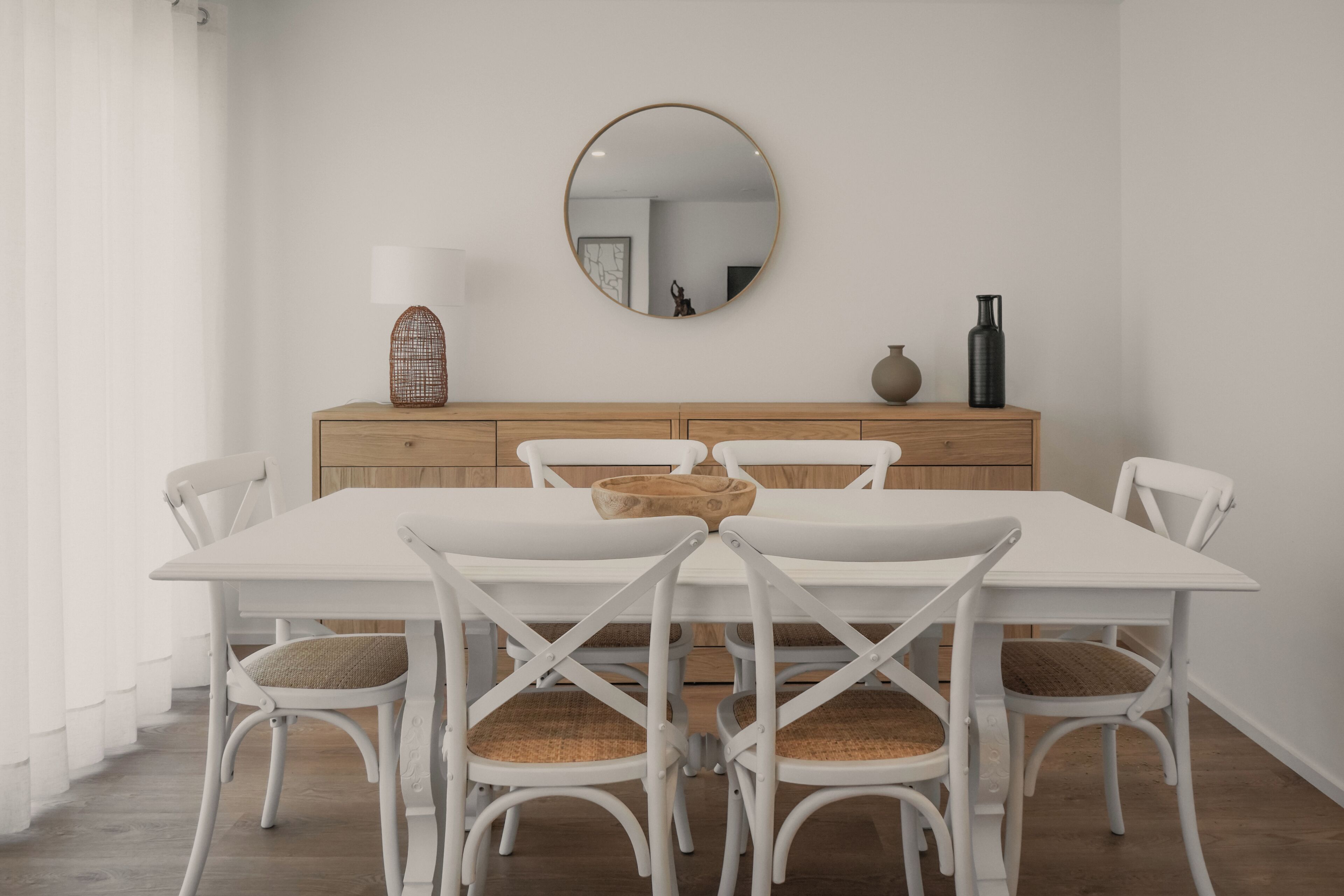 A dining area with a white table, wicker chairs, a wooden cabinet, and a round mirror on the wall.