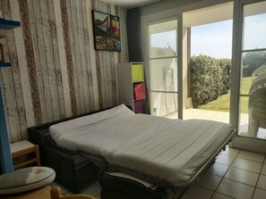 A bedroom with a bed, a chair, a small table, a window, and a view of the outdoors. - T1 cabine pour 4 avec terrasse piscine l'été (ARZON)