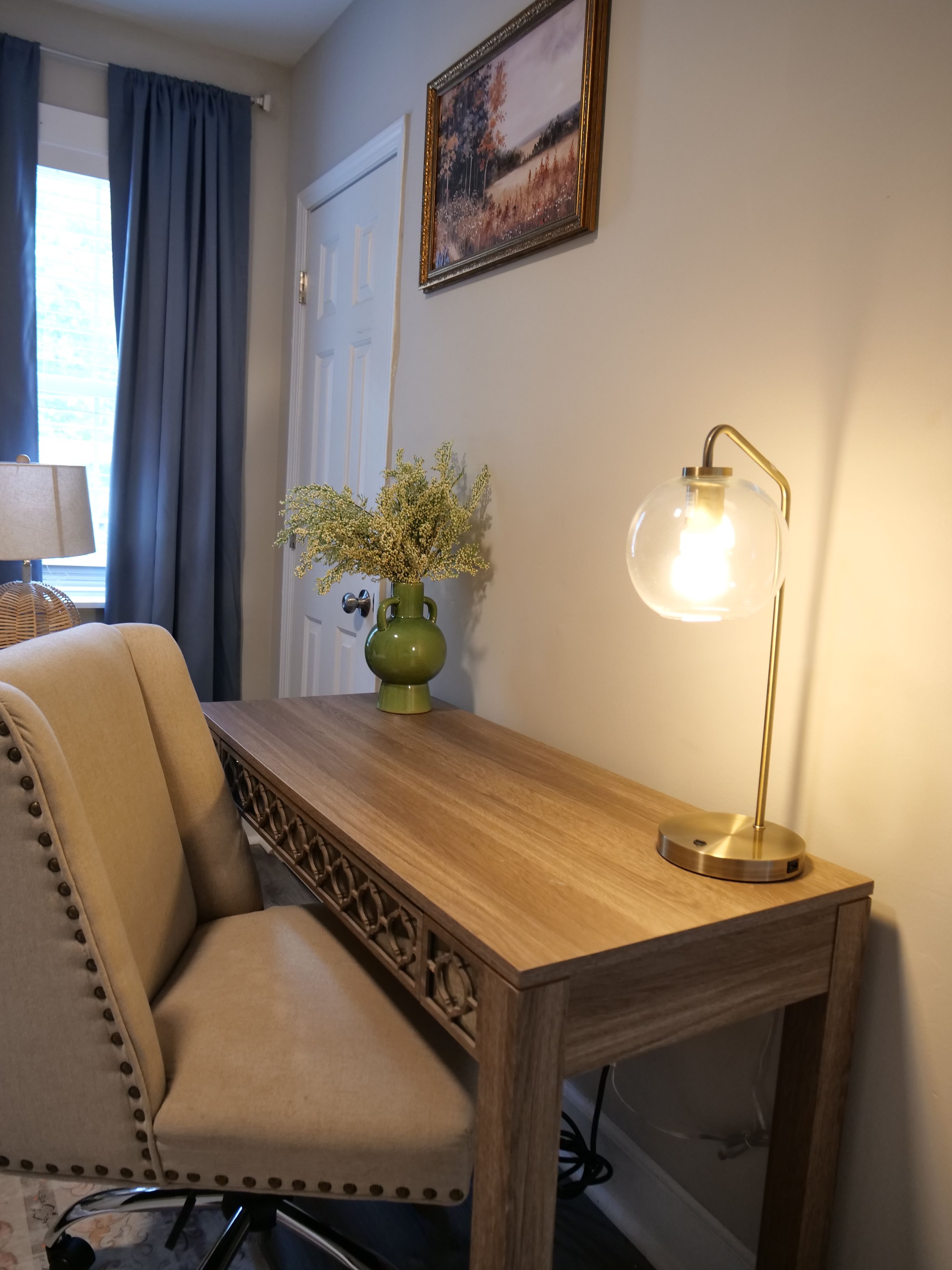 Una habitación con una mesa de madera, un jarrón verde con flores, una lámpara y una silla.