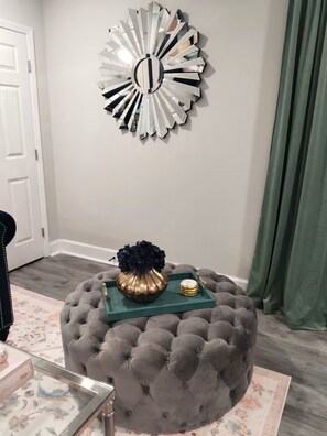 Ein Fussbank mit Polsterung, eine dekorative Vase und ein Tablett, ein Spiegel an der Wand und ein grüner Vorhang.