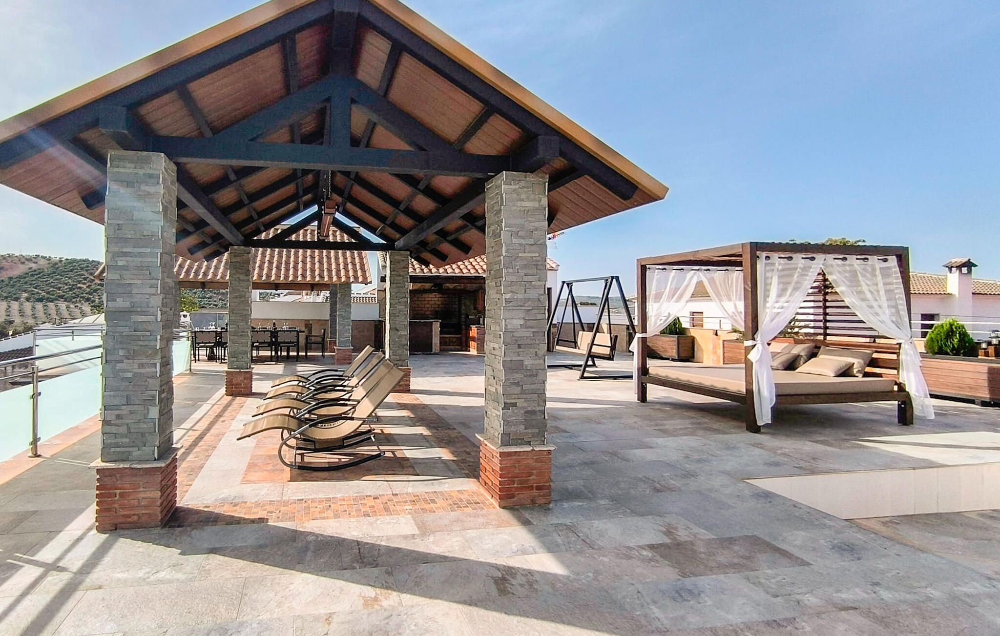 A covered outdoor seating area with stone pillars, a pergola with a swing set, and a view of a building in the background.