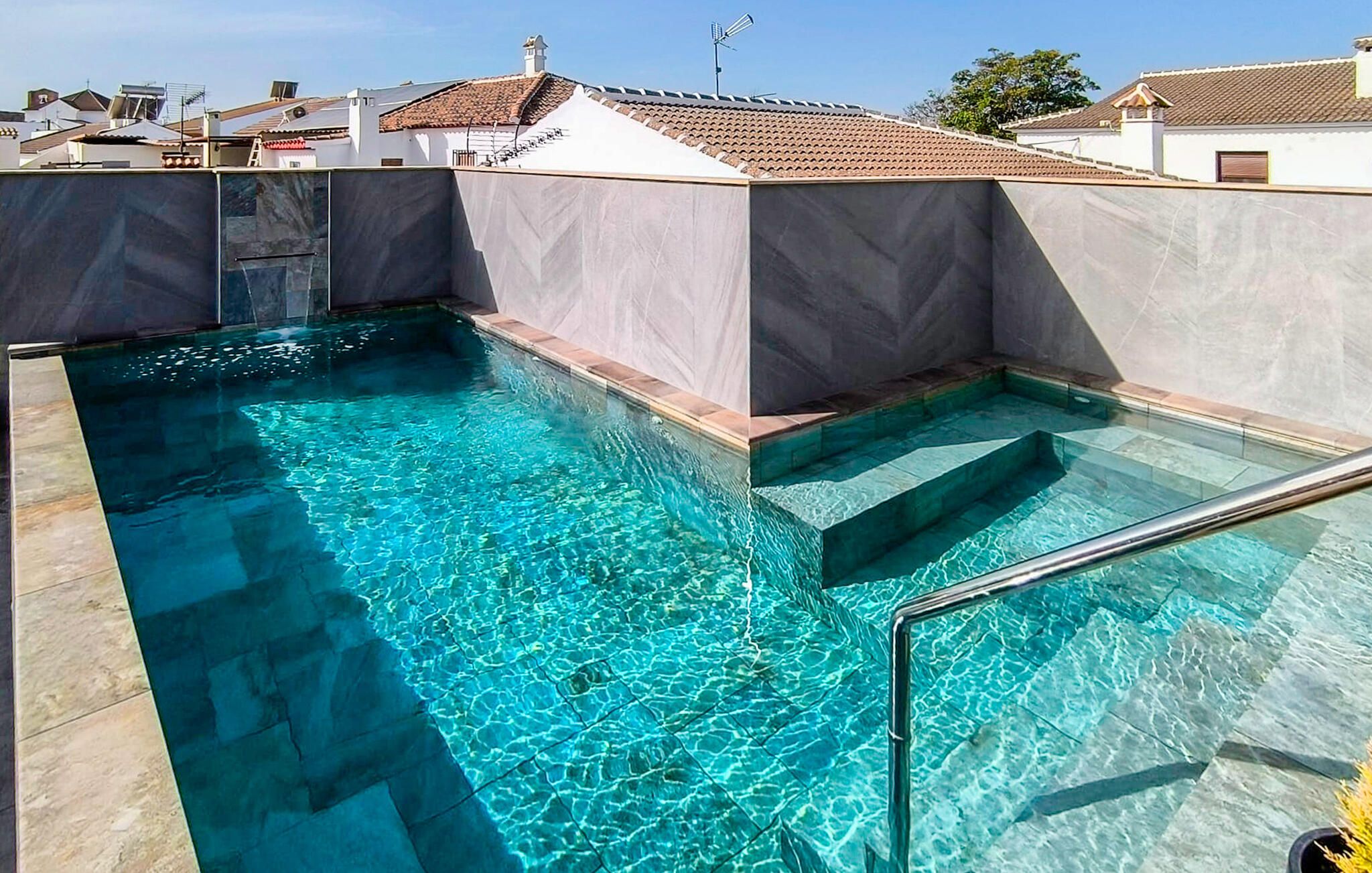 Uma piscina com água azul cristalina, rodeada por um deck ladrilhado e um muro de pedra.