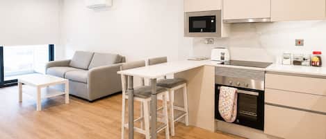 Een moderne keuken met een magnetron, broodrooster en oven, een eethoek met een tafel en stoelen, en een bank in een lichte kamer met houten vloeren.