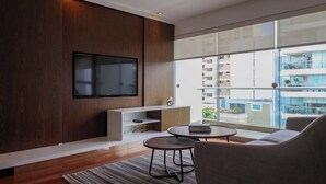 Ein modernes Wohnzimmer mit einem Flachbildfernseher, einer Couch, einem Couchtisch und Blick auf ein Gebäude durch große Fenster.