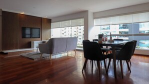 Ein modernes Wohnzimmer mit Fernseher, einer Couch, einem Esstisch mit Stühlen und großen Fenstern mit Blick auf ein Gebäude.