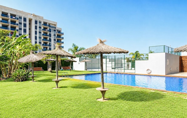 A swimming pool with grass and thatched umbrellas, a modern building in the background, and a clear sky.