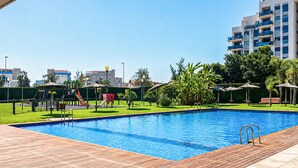 Ein Schwimmbecken mit Holzterrasse, umgeben von Grünpflanzen und einem Spielplatz mit Rutschen.