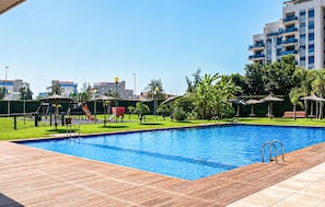 A swimming pool with a wooden deck, surrounded by greenery and a playground with slides.
