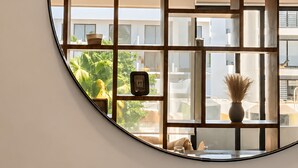 A circular mirror reflecting a room with a ceiling fan, a window with a view of greenery, and a vase on a table.