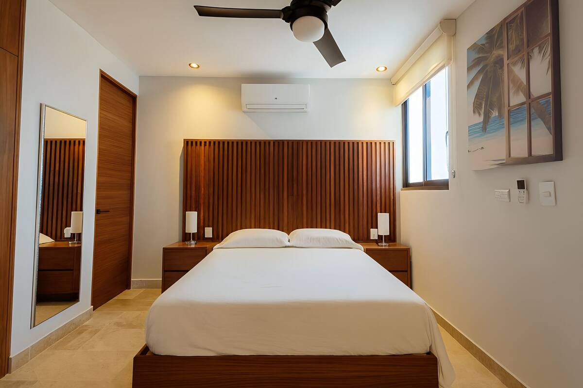 Habitación de hotel moderna con una cama grande, cabecera de madera, ventilador de techo, equipo de aire acondicionado, lámparas de noche y una ventana con vistas.