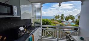 Ein Balkon mit Blick auf das Meer und Palmen, eine Küche mit Mikrowelle sowie ein Poolbereich.