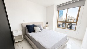 Ein modernes Schlafzimmer mit einem großen Bett, einem Nachttisch, einer Lampe und Blick auf die Stadt durch ein großes Fenster.