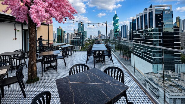 Ein Sitzbereich im Freien mit Tischen und Stühlen, ein Baum mit rosa Blüten und Blick auf die Stadt.