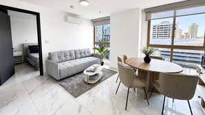 Ein modernes Wohnzimmer mit einer Couch, einem Esstisch, Stühlen, einer Zimmerpflanze und Blick auf die Stadt durch große Fenster.